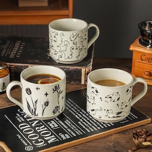 May include: Three ceramic coffee mugs with distinctive designs. One mug has abstract faces, another has floral patterns, and the third shows cat illustrations. The mugs contain coffee and tea, and are on a wooden surface.