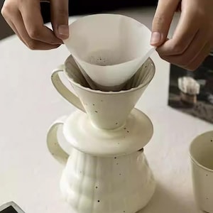 May include: Cream-colored ceramic pour-over coffee maker with a matching lid and a handle. A white paper filter is being placed into the top cone. The coffee maker has a speckled design and is sitting on a white surface.