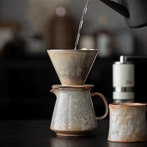 May include: A ceramic pour-over coffee set with a cone-shaped filter and a matching carafe. The set is in shades of brown and gray, with water pouring into the filter. A mug is in the background.