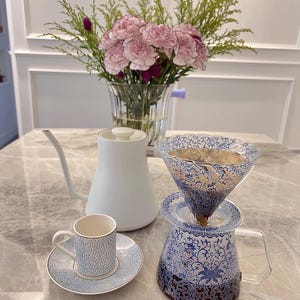 May include: A white gooseneck kettle, a small cup and saucer set, and a coffee dripper with a blue floral design sit on a marble surface. A bouquet of pink flowers in a glass vase is in the background. The coffee dripper is in use.