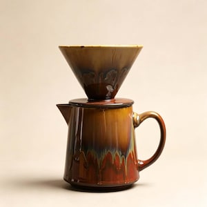 Könnte beinhalten: Ein Keramik-Kaffeefilteraufsatz, der auf einer passenden Keramikkanne steht. Beide Teile haben eine glänzende, mehrfarbige Glasur mit vermischten Braun-, Bernstein- und Dunkelrottönen, die ein markantes, tropfendes Finish aufweisen.
