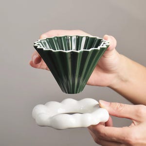 Könnte beinhalten: Ein dunkelgrüner Keramik-Kaffeefilter mit gerilltem Design und weißem Rand, gehalten über einem weißen Keramiksockel. Der Filter ist kegelförmig, und der Sockel ist ein Ring mit gewelltem Rand. Der Hintergrund ist ein neutrales Grau.
