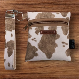 May include: A white and brown cowhide-patterned wristlet and matching pouch with a brown zipper and a small leather tag that says "Handmade". The wristlet has a silver clasp and key ring.