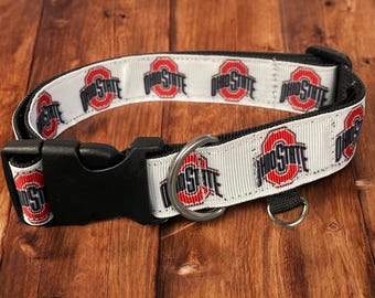 May include: White dog collar with the Ohio State University logo in red and grey. The collar has a black buckle and a metal D-ring for leash attachment. The Ohio State logo is repeated along the white collar.
