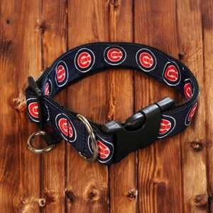 Op de afbeelding: Een marineblauwe hondenriem met het Chicago Cubs-logo in rood en wit. De halsband heeft een zwarte plastic gesp en een metalen D-ring voor het bevestigen van de lijn. De halsband wordt gepresenteerd op een houten oppervlak.