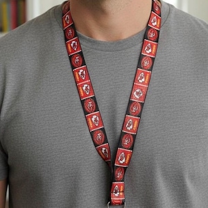 May include: A red and black lanyard featuring a repeating pattern of a team logo. The lanyard is worn around the neck and has a metal clasp at the bottom. The person in the image is wearing a gray t-shirt.