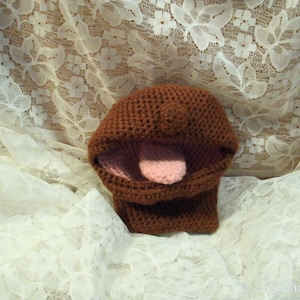May include: A handmade brown crocheted puppet with a pink tongue. The puppet is open-mouthed, with a rounded top and a rectangular base. The background is a white lace fabric.