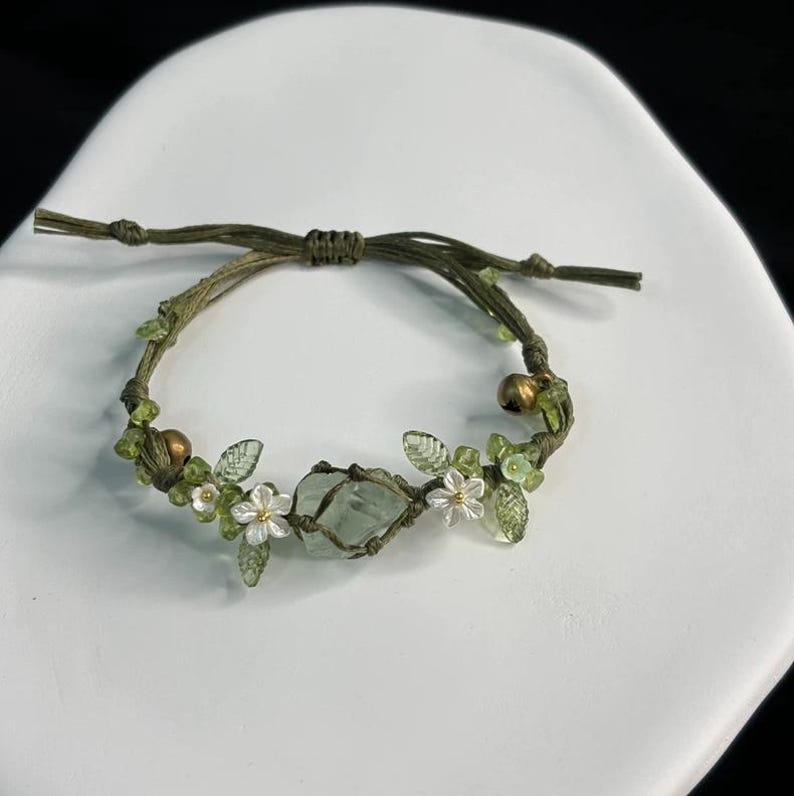 Pu&ograve; includere: Braccialetto fatto a mano con una pietra centrale azzurra, ornato da piccoli fiori bianchi, foglie verdi e perline. Il braccialetto in cordoncino verde oliva include una piccola campana color oro. Il braccialetto &egrave; progettato con un nodo macram&egrave;.