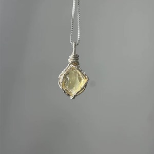 May include: A necklace featuring a pale yellow, translucent, raw gemstone pendant encased in a silver-colored wire-wrapped cage. The pendant hangs from a delicate silver-colored chain that connects to the top of the wire-wrapped setting.