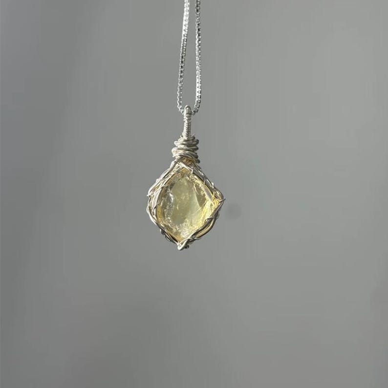 May include: A necklace featuring a pale yellow, translucent, raw gemstone pendant encased in a silver-colored wire-wrapped cage. The pendant hangs from a delicate silver-colored chain that connects to the top of the wire-wrapped setting.