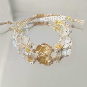 Pode incluir: Uma pulseira de macramé feita à mão com cristais transparentes e amarelo dourado, pequenas flores brancas e um cordão tecido. A pulseira tem uma estética boêmia delicada. O texto "@LITTLE RED" é visível.