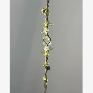 Pu&ograve; includere: Una decorazione da appendere in macram&egrave; fatta a mano con un cristallo verde chiaro, fiori bianchi e perline verdi. Il design incorpora piccole campane color oro e delicate foglie verdi, creando un'estetica naturale e terrosa.