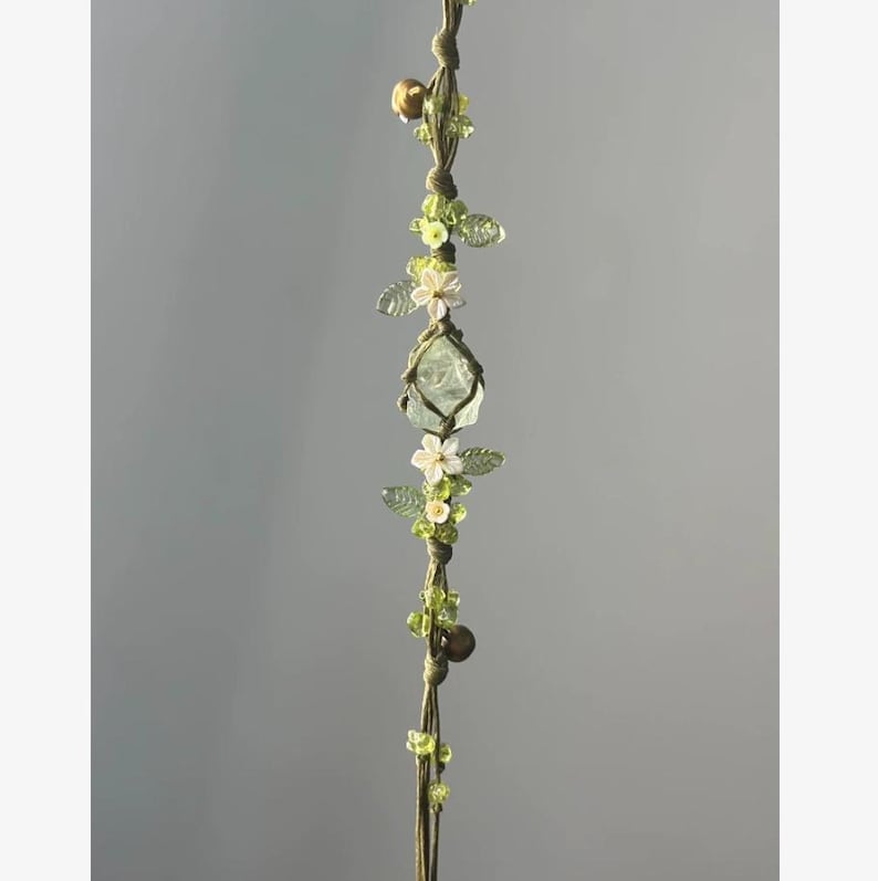 Pu&ograve; includere: Una decorazione da appendere in macram&egrave; fatta a mano con un cristallo verde chiaro, fiori bianchi e perline verdi. Il design incorpora piccole campane color oro e delicate foglie verdi, creando un'estetica naturale e terrosa.