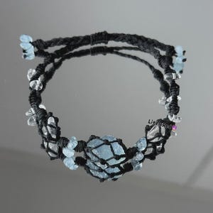 May include: A macrame bracelet featuring a large, light blue crystal at the center, surrounded by smaller clear and light blue crystals. The bracelet is crafted with black cord, creating a striking contrast with the gemstones. The design is symmetrical and elegant.