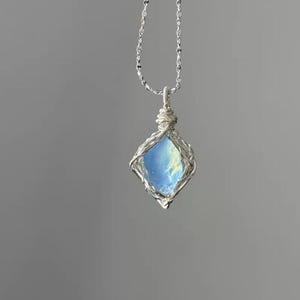 May include: A silver wire-wrapped pendant featuring a translucent, light blue, diamond-shaped stone with internal cloudy inclusions. The pendant hangs from a delicate silver chain, showcasing intricate metalwork that secures the gemstone.