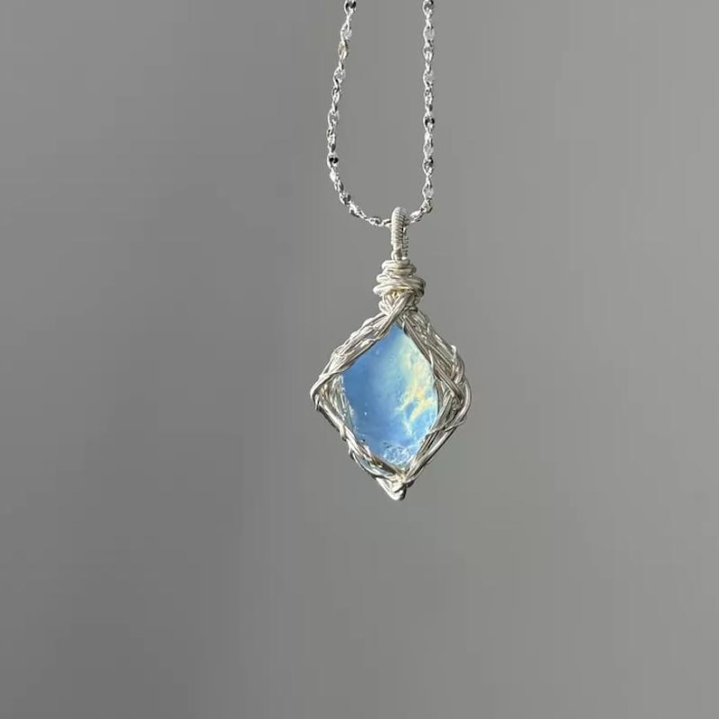 May include: A silver wire-wrapped pendant featuring a translucent, light blue, diamond-shaped stone with internal cloudy inclusions. The pendant hangs from a delicate silver chain, showcasing intricate metalwork that secures the gemstone.