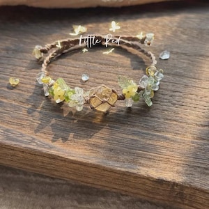 May include: A handmade bracelet featuring a central citrine crystal, surrounded by small yellow and green beads, and delicate floral accents. The bracelet is crafted with a brown cord and has the text "Little Red".