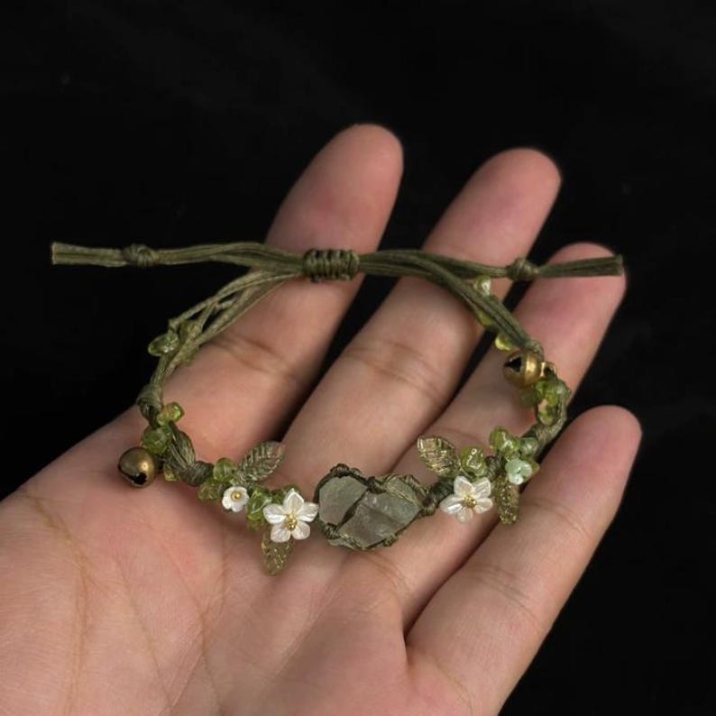 Pu&ograve; includere: Un braccialetto verde regolabile con una pietra centrale verde chiaro di forma irregolare. Il braccialetto &egrave; decorato con piccoli fiori bianchi, foglie verdi e minuscole campanelle color oro. Il braccialetto &egrave; tenuto in una mano su sfondo scuro.