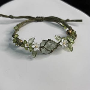 Pu&ograve; includere: Bracciale in macram&egrave; verde fatto a mano con un cristallo verde chiaro al centro. Il bracciale &egrave; decorato con piccoli fiori bianchi, foglie verdi e perline verdi. Questo bracciale regolabile &egrave; un gioiello unico.
