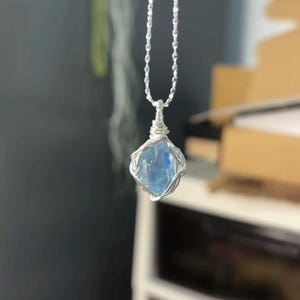 May include: A necklace featuring a raw, light blue, translucent gemstone pendant encased in a silver-colored wire wrap. The pendant hangs from a delicate silver-colored ball chain, with the wire coiled tightly at the top to secure the stone.