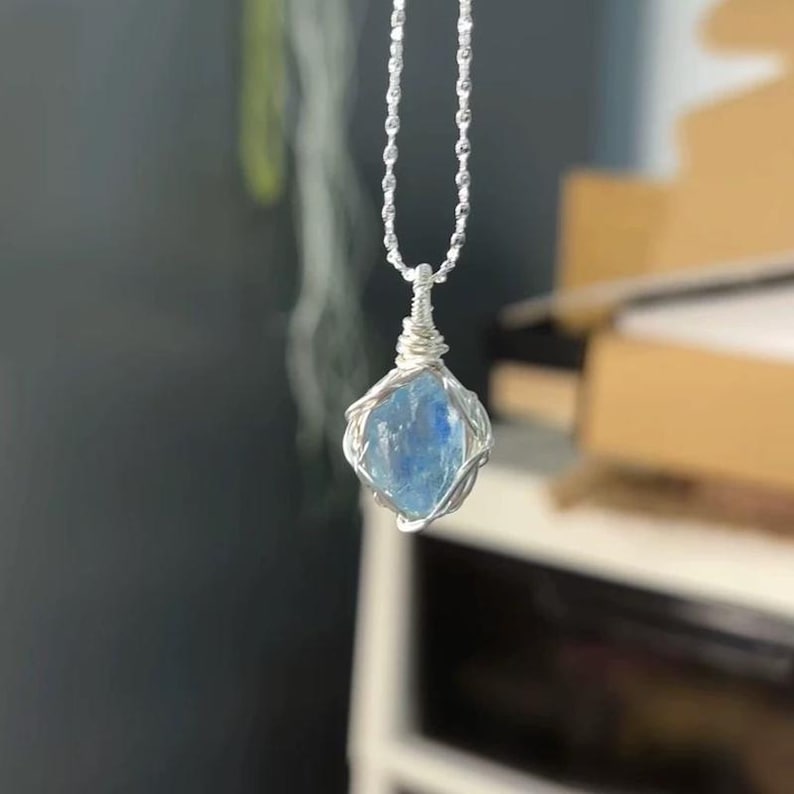 May include: A necklace featuring a raw, light blue, translucent gemstone pendant encased in a silver-colored wire wrap. The pendant hangs from a delicate silver-colored ball chain, with the wire coiled tightly at the top to secure the stone.