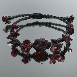 Puede incluir: Pulsera de macramé negro hecha a mano adornada con piedras de granate rojo oscuro y detalles florales. La pulsera presenta flores de granate y otras formas, con una longitud total de aproximadamente 18 cm.