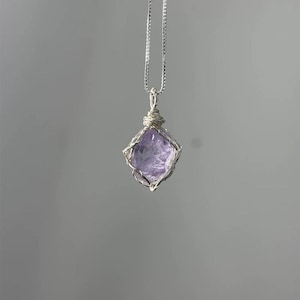 May include: A silver-toned necklace featuring a raw, faceted light purple gemstone pendant encased in a delicate wire-wrapped cage. The pendant hangs from a thin, matching silver-toned chain, showcasing the natural texture of the stone.