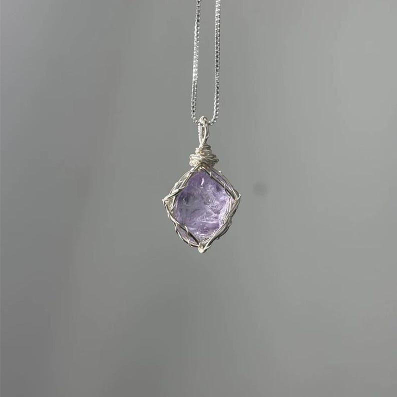 May include: A silver-toned necklace featuring a raw, faceted light purple gemstone pendant encased in a delicate wire-wrapped cage. The pendant hangs from a thin, matching silver-toned chain, showcasing the natural texture of the stone.