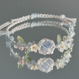 以下が含まれることがあります： 花と宝石のアクセントが付いた、2つのライトブルーのマクラメブレスレット。各ブレスレットは、白い花、緑の葉、小さな青い石に囲まれた、中央の青い宝石が特徴です。ブレスレットは編み込みコードとタッセルの端が付いています。