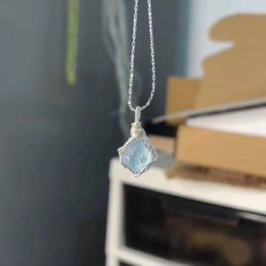 May include: A silver-toned chain necklace featuring a pendant made of a single, raw, light blue gemstone. The stone is secured by a delicate silver-colored wire wrapping that encircles the gem and extends upward to connect to the thin metal chain.