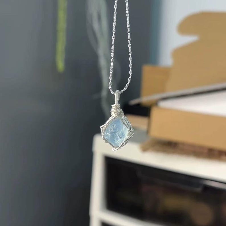 May include: A silver-toned chain necklace featuring a pendant made of a single, raw, light blue gemstone. The stone is secured by a delicate silver-colored wire wrapping that encircles the gem and extends upward to connect to the thin metal chain.