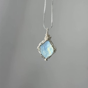 May include: A pendant necklace featuring a raw, light blue, translucent stone encased in a silver-colored wire-wrapped setting. The stone has a diamond-like shape and is suspended from a thin, silver-colored box chain that hangs vertically.