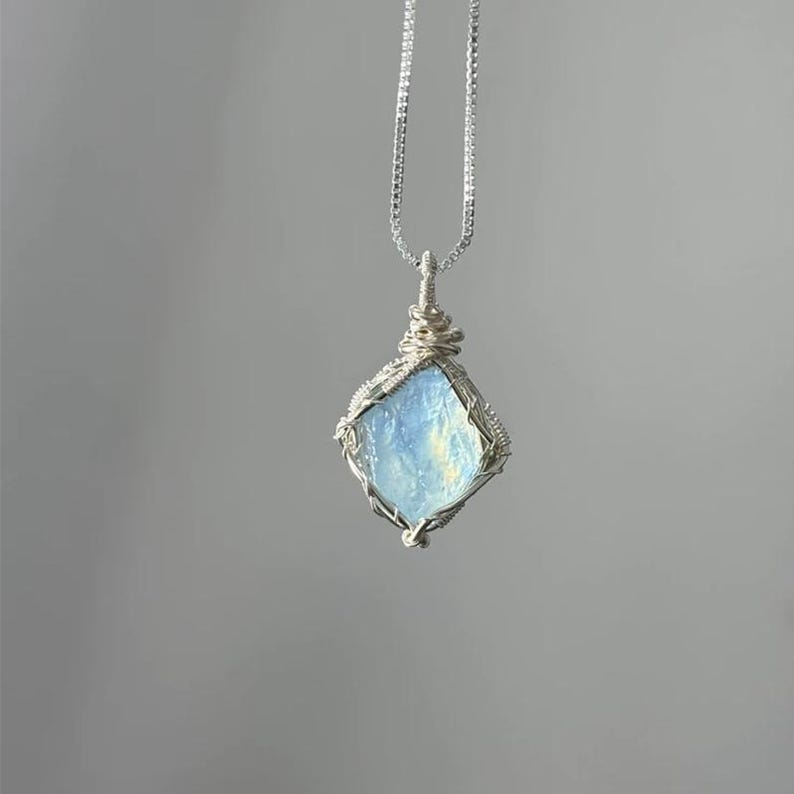 May include: A pendant necklace featuring a raw, light blue, translucent stone encased in a silver-colored wire-wrapped setting. The stone has a diamond-like shape and is suspended from a thin, silver-colored box chain that hangs vertically.