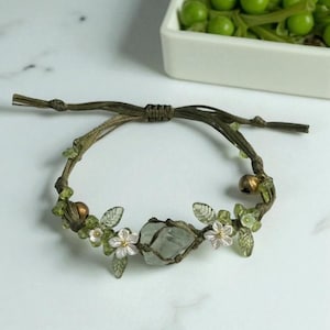 Pu&ograve; includere: Braccialetto fatto a mano con una pietra centrale chiara avvolta in macram&egrave;. Decorato con piccole perline verdi, delicati fiori bianchi e dettagli di foglie trasparenti, il tutto su un cordoncino marrone. Circonferenza di circa 18 cm.
