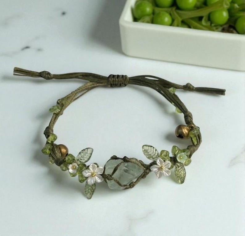 Pu&ograve; includere: Braccialetto fatto a mano con una pietra centrale chiara avvolta in macram&egrave;. Decorato con piccole perline verdi, delicati fiori bianchi e dettagli di foglie trasparenti, il tutto su un cordoncino marrone. Circonferenza di circa 18 cm.