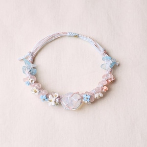 May include: A bracelet featuring a central clear crystal wrapped in pink cord, accented with light blue and pink beads, small white and blue flower charms, and light blue stone chips, all strung on a braided light pink and white cord band.