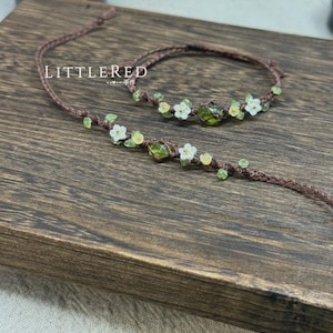 May include: A brown macrame necklace and bracelet set. The jewelry is adorned with small white flowers and green beads. The word "LITTERED" is printed on the wooden surface. The jewelry is displayed on a wooden surface.