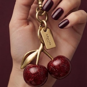 Peut inclure: Un porte-clés en métal doré composé de deux breloques cerises rondes et pailletées rouge foncé, fixées à une tige avec une feuille. Le porte-clés comprend un anneau circulaire et une petite étiquette rectangulaire gravée du nom COACH.