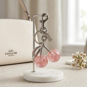 以下が含まれることがあります： ピンクのグリッターが詰まった球体、シルバーの茎、葉が付いた、コーチのチェリー型のキーホルダー。 キーホルダーはシルバーのクリップと小さなタグに付いています。 キーホルダーは白い大理石のスタンドに展示されています。