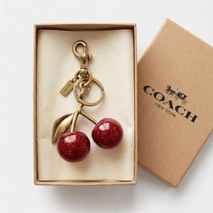 Peut inclure: Un porte-clés en forme de cerises de la marque Coach, composé de deux cerises rouges pailletées fixées à une tige, une feuille et un mousqueton en métal doré. Le porte-clés est présenté dans une boîte en carton beige avec le logo Coach New York.