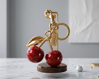 May include: A gold-toned metal cherry keyring featuring two glossy red spheres, a gold leaf accent, a circular ring, a lobster clasp, and a small rectangular tag engraved with the word COACH. The keyring is displayed on a dark wood base.