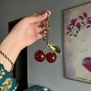 以下が含まれることがあります： 金色のキーホルダー。赤色のキラキラしたチェリーが2つ、金色の葉と茎が付いています。キーホルダーにはロブスタークラスプとチェーンリンクが付いています。チェリーの直径は約2.5cmです。