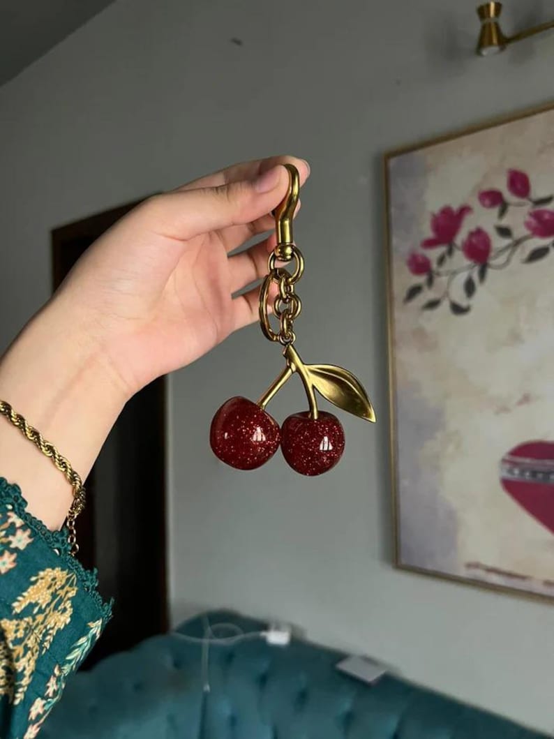 以下が含まれることがあります： 金色のキーホルダー。赤色のキラキラしたチェリーが2つ、金色の葉と茎が付いています。キーホルダーにはロブスタークラスプとチェーンリンクが付いています。チェリーの直径は約2.5cmです。