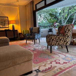 May include: A living room scene featuring two patterned armchairs, a large ottoman, and a colorful rug. The rug has a pink and cream background with intricate designs. The room has large windows and a view of greenery.