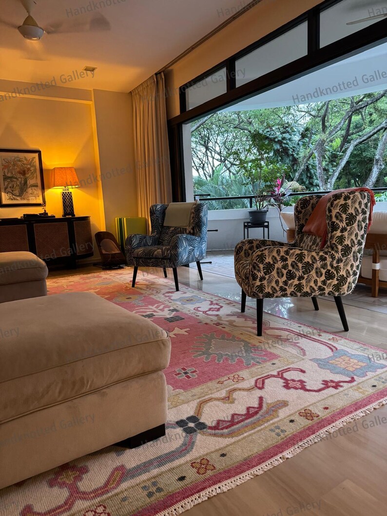May include: A living room scene featuring two patterned armchairs, a large ottoman, and a colorful rug. The rug has a pink and cream background with intricate designs. The room has large windows and a view of greenery.