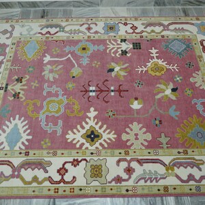 May include: A rectangular rug with a pink central field and a cream-colored border. The rug features intricate floral and geometric patterns in shades of blue, yellow, and green. The rug is placed on a gray tiled floor.
