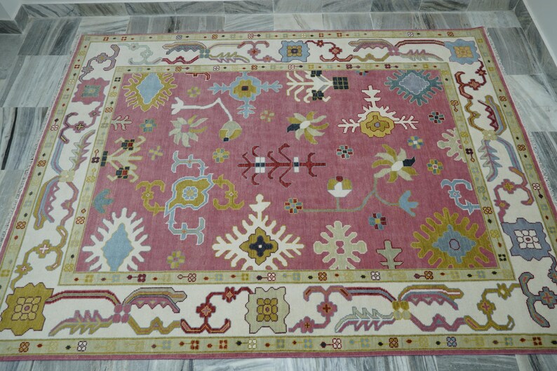May include: A rectangular rug with a pink central field and a cream-colored border. The rug features intricate floral and geometric patterns in shades of blue, yellow, and green. The rug is placed on a gray tiled floor.