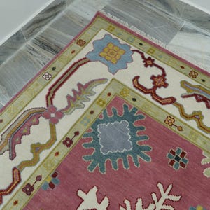 May include: A section of a decorative rug with a pink field and a cream-colored border. The border features floral and geometric patterns in shades of blue, green, and red. The rug is placed on a marble tile floor.
