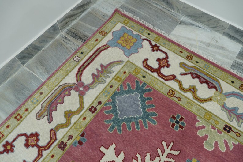 May include: A section of a decorative rug with a pink field and a cream-colored border. The border features floral and geometric patterns in shades of blue, green, and red. The rug is placed on a marble tile floor.