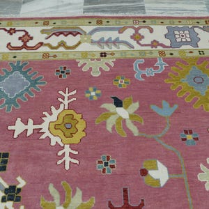 May include: A detailed shot of a decorative rug with a pink central field. The rug features a complex floral pattern in white, yellow, blue, and red, with a cream-colored border. The design is symmetrical and intricate.